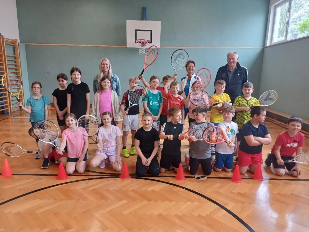 Eine Gruppe von Kindern und zwei Erwachsenen posiert mit Tennisschlägern in einem Gymnasium für ein Foto.
