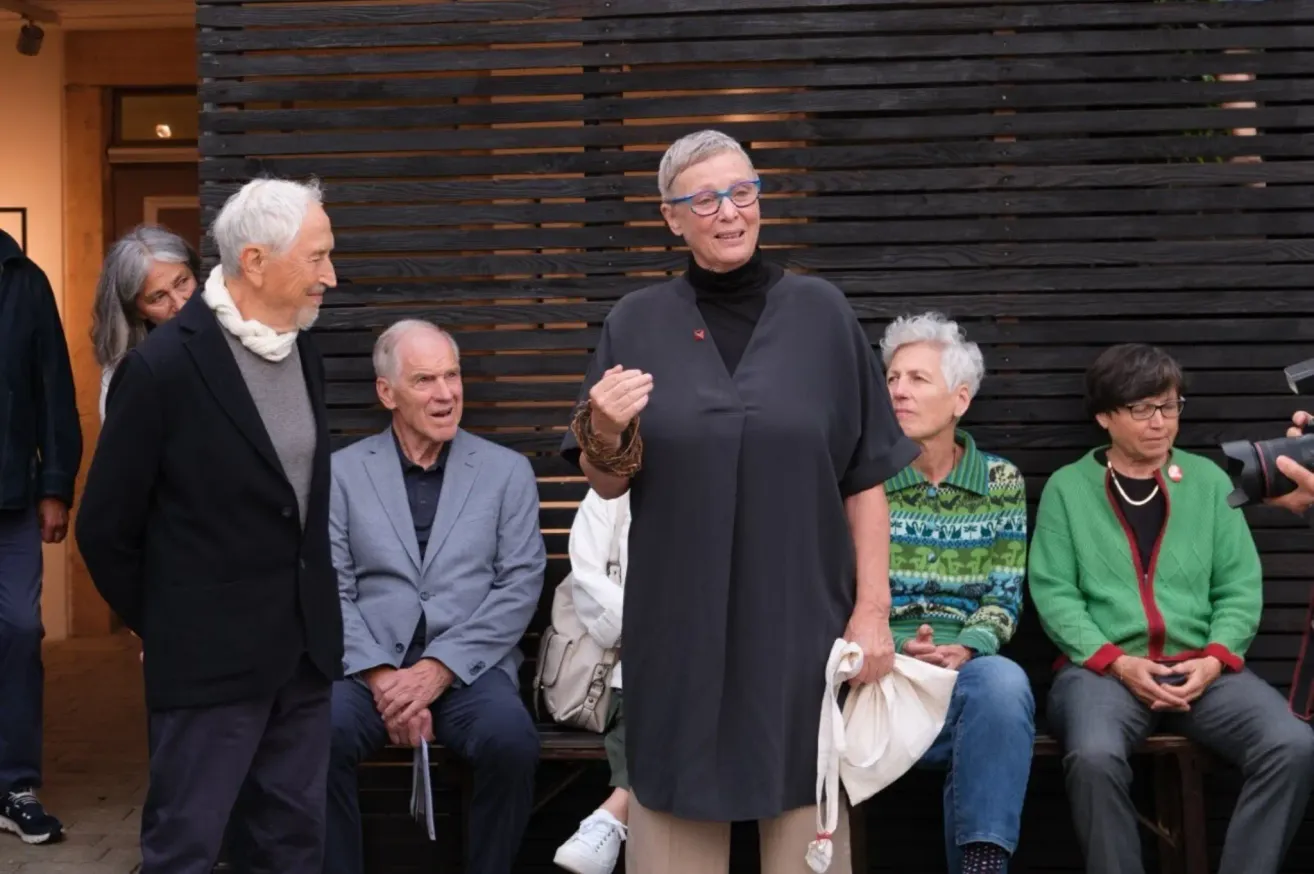 An elderly woman stands between two men. She wears glasses and a dark blouse. The man on the left wears a scarf and jacket. The others are seated and look at the camera.