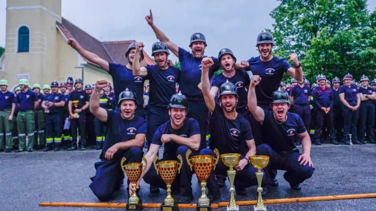 Eine Gruppe von Feuerwehrleuten in Helmen und blauen Uniformen posiert mit ihren Trophäen und feiert einen Sieg. Sie sind von anderen Menschen umgeben, von denen einige ebenfalls Helme tragen.