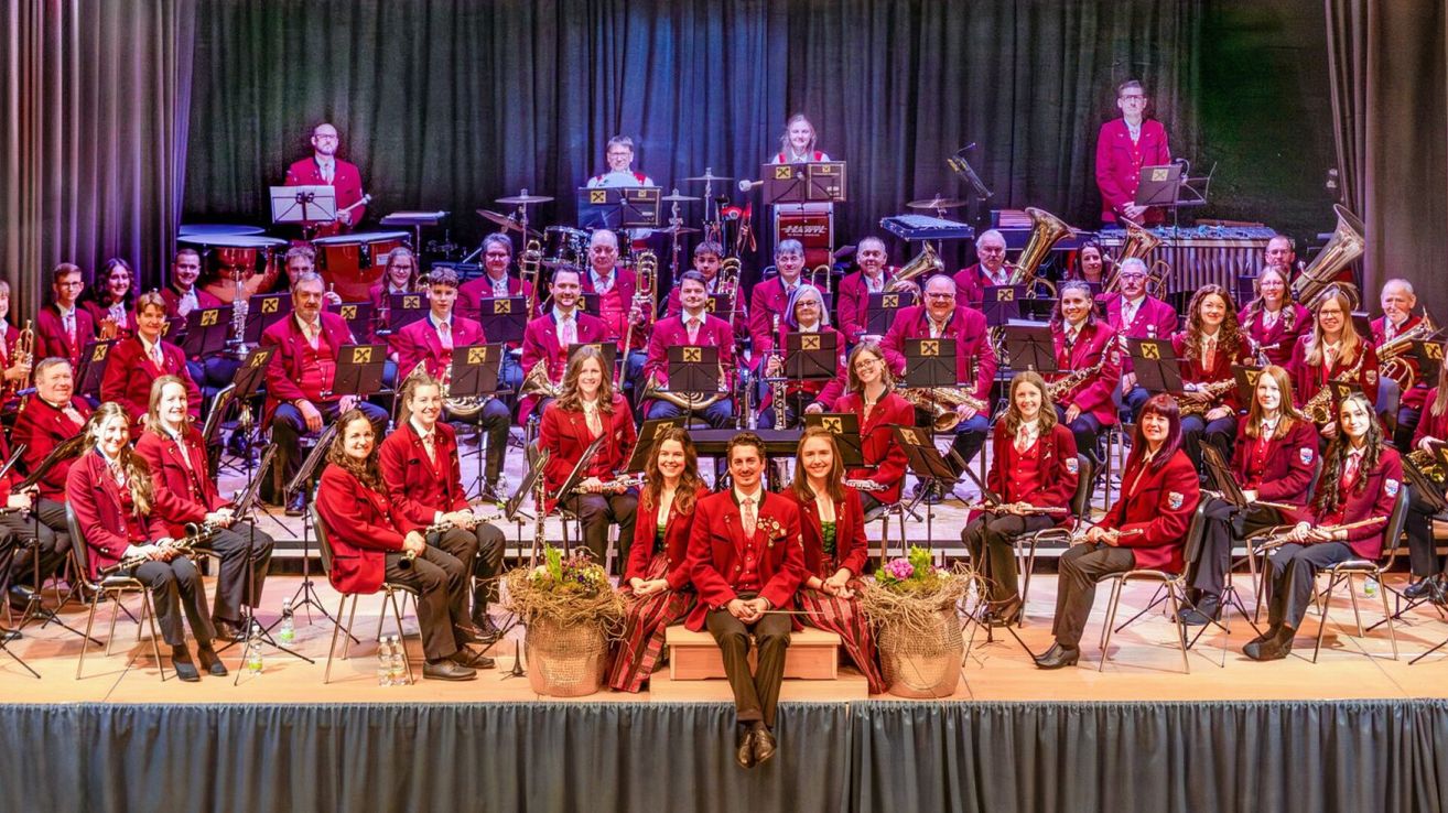 A band on stage, all dressed in red with some in front holding instruments. A group of people sitting on chairs and some standing behind them.