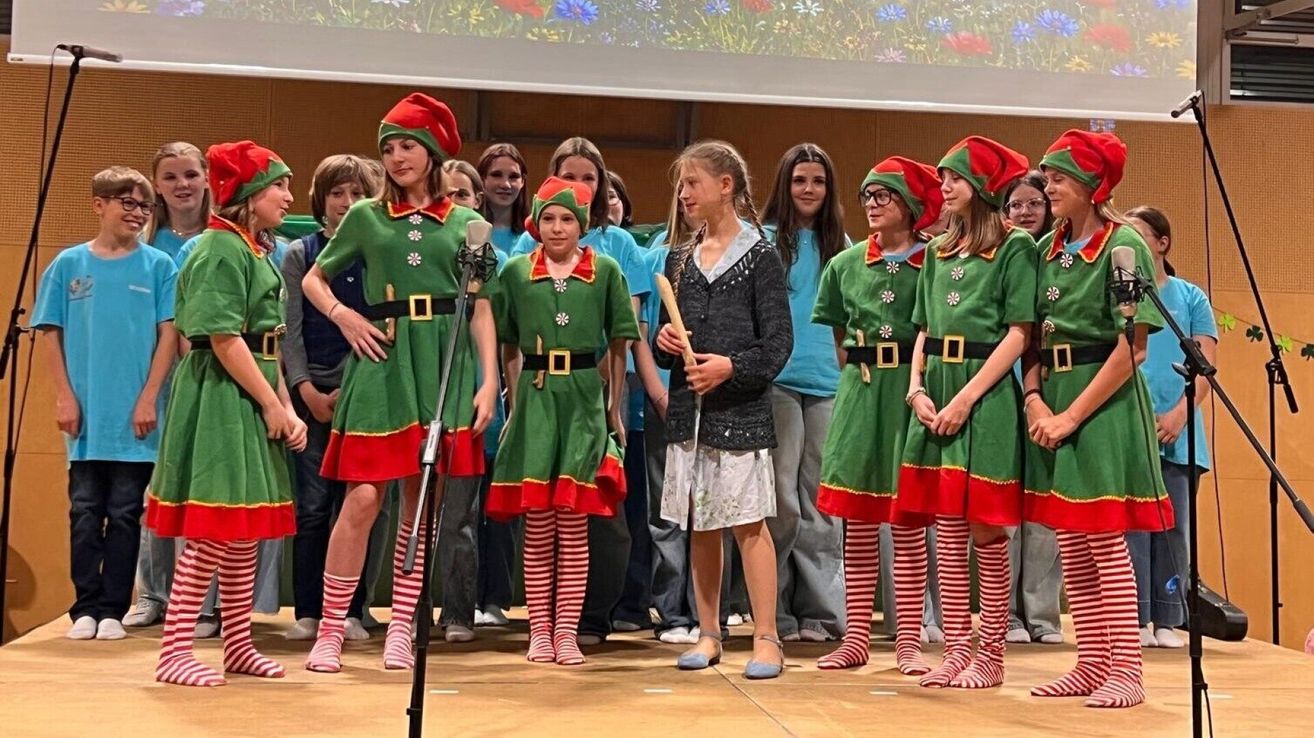 A group of children dressed as elves are performing on stage, some holding props. Behind them, more children and adults are standing.
