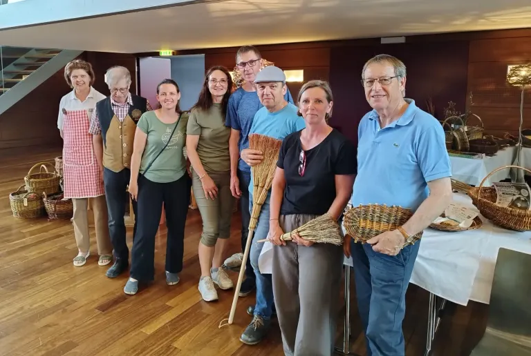 A group of people stand together, smiling. Some hold woven brooms. They are indoors, with wooden floors and a white table in the background.