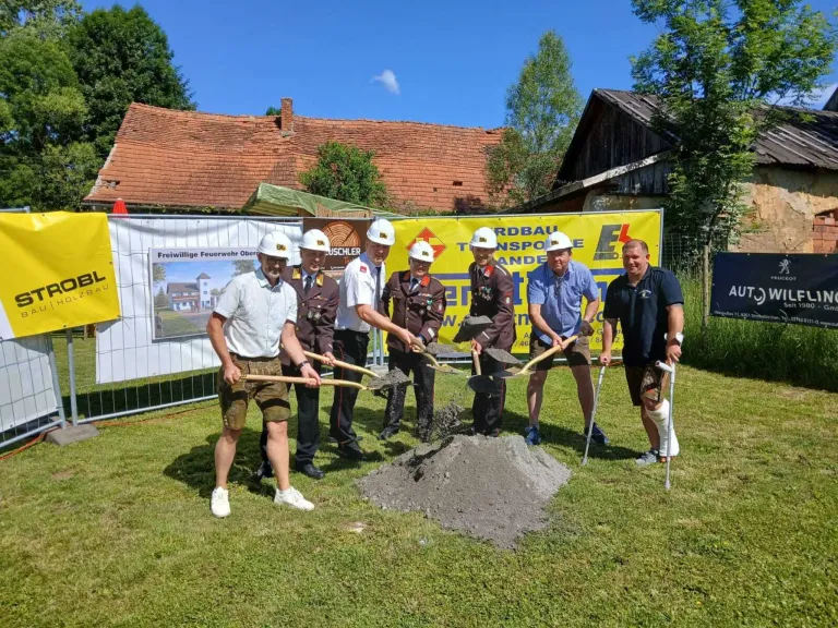 Sieben Männer in harten Arbeitshelmen stehen um einen Haufen Erde herum und halten Schaufeln. Zwei Männer haben Gehstützen an den Beinen. Dahinter wird ein gelbes Banner gezeigt.