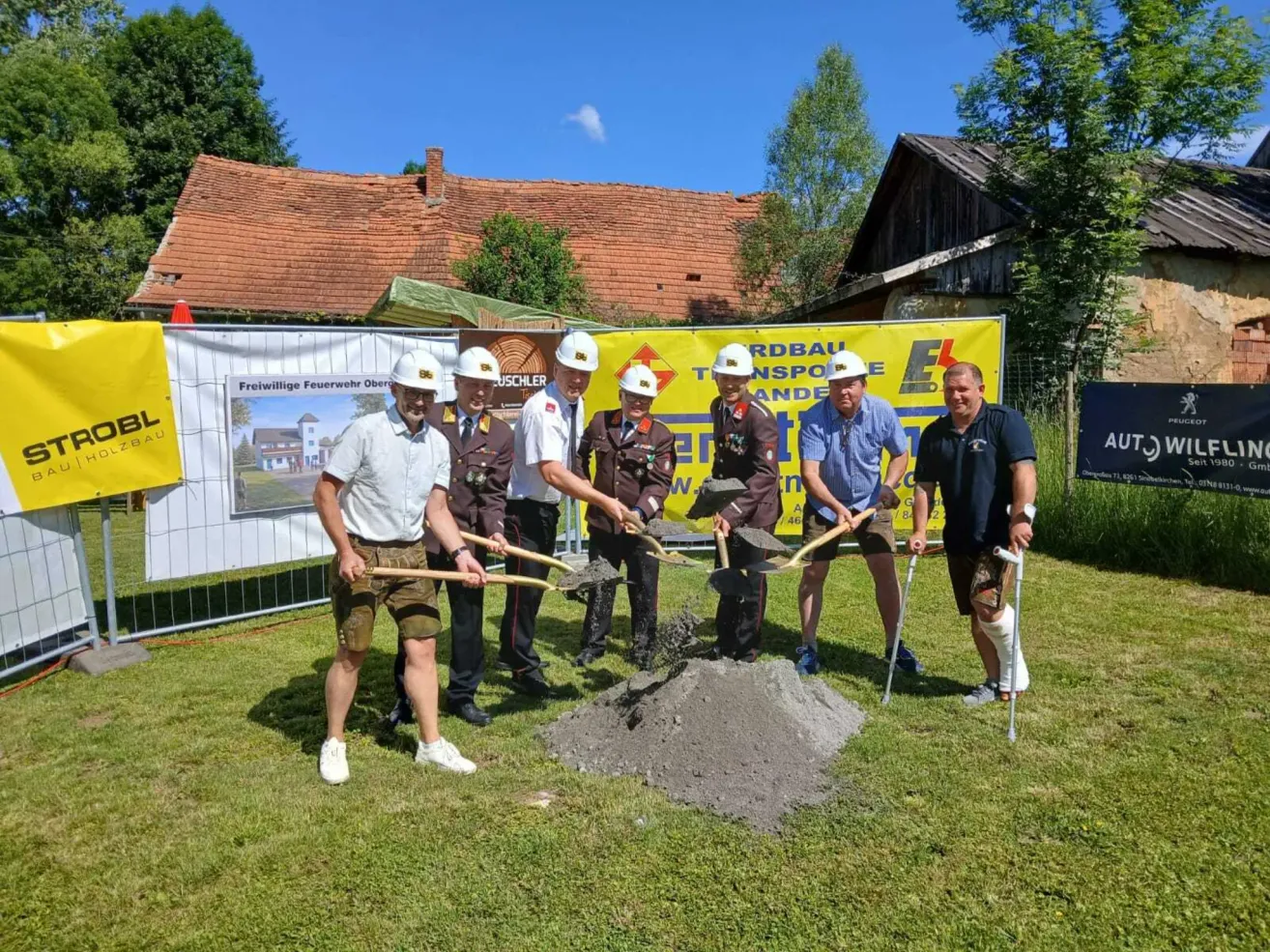 Seven men in hard hats stand around a pile of dirt, holding shovels. Two men have walking casts on their legs. Behind them, a yellow banner is displayed.