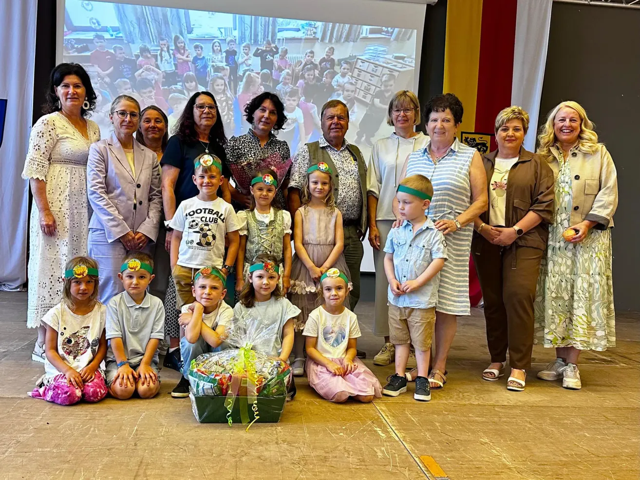 A group of adults and children, some wearing headbands, are posing for a photo on a stage with a backdrop. Some children sit on the floor with a basket in front of them.