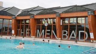 An outdoor pool with several people swimming. The pool is surrounded by a building with large windows, a clock, and the word 'MEN' prominently displayed.