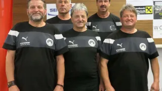 Five men wearing black sports jerseys with a white stripe and the word Wintersport on the front are standing in front of a wall with the number 00 on it.