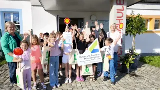 Children holding bags and a certificate stand in front of a building labeled 'Klima- und Energie-Modellregionen.' They are all smiling and raising their hands. The woman in the middle holds a sign.