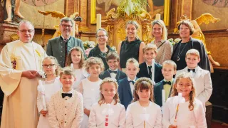 A group of children and adults in white dresses stand in a church, holding candles. A priest and adults stand behind them. An altar is decorated with candles, flowers, and a statue.