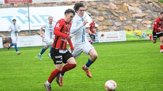 Soccer players in jerseys are running on a field. One is wearing red, and another is in white. They are chasing a soccer ball.
