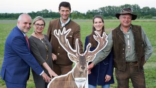A group of individuals stand in a field with a cardboard cutout of a deer, dressed in traditional German attire, smiling and posing for a photo.