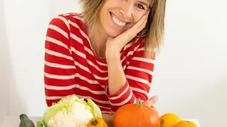 A smiling woman poses with a table of various fruits and vegetables. She wears a red and white striped shirt and has her hand under her chin.