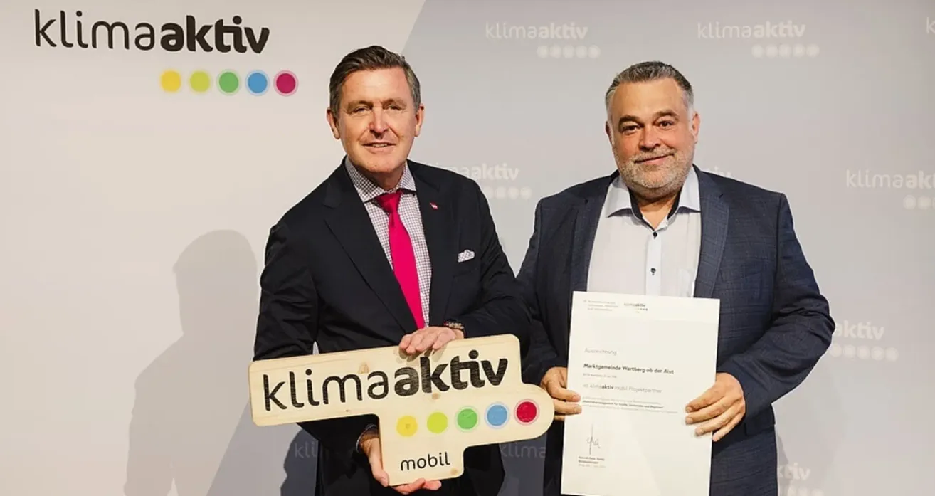 Zwei Männer in Anzügen stehen zusammen, einer hält eine Urkunde und der andere ein Holzschild mit dem Wort 'klimaaktiv' darauf. Der Mann im Anzug auf der rechten Seite lächelt und hält eine Urkunde. Der Mann auf der linken Seite trägt eine rosa Krawatte. Hinter ihnen ist eine weiße Wand mit Logos.