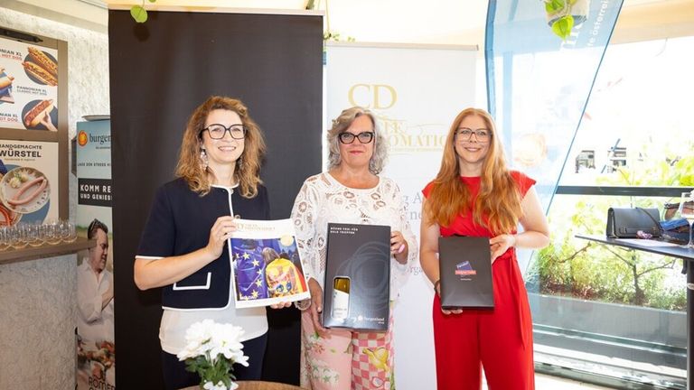 Drei Frauen stehen zusammen und halten Bücher und Boxen. Eine hat eine Zeitschrift mit buntem Cover, eine andere eine Weinflasche und die dritte hält eine schwarze Box. Sie lächeln und posieren für die Kamera.