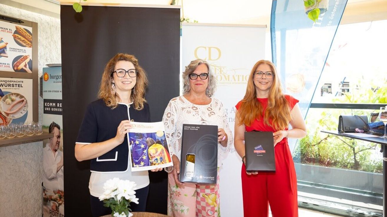 Drei Frauen stehen zusammen und halten Bücher und Boxen. Eine hat eine Zeitschrift mit buntem Cover, eine andere eine Weinflasche und die dritte hält eine schwarze Box. Sie lächeln und posieren für die Kamera.