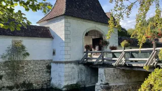 A stone building with a brown roof and a wooden bridge over a river. A potted plant is on the bridge.