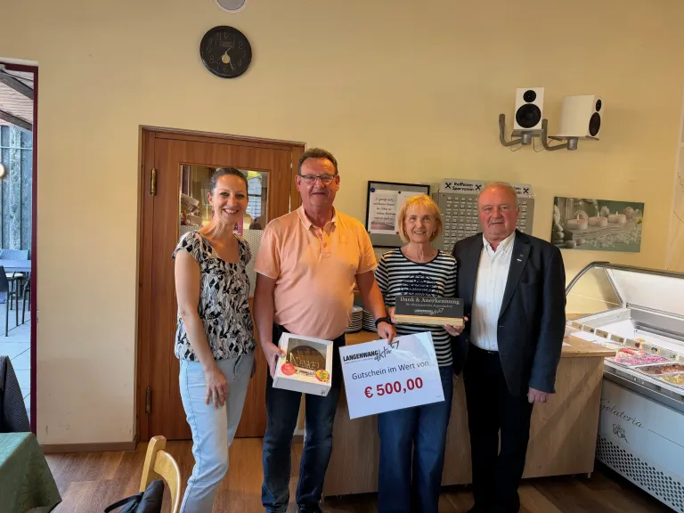 Four people stand inside a room holding a plaque and a voucher. The woman on the left holds a box of cake. A clock and speakers are on the wall.