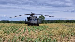 Ein Militärhubschrauber mit offenen Türen steht in einem trockenen Feld. Bäume und ein bewölkter Himmel sind im Hintergrund.