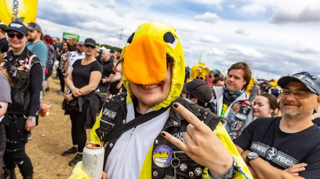 Ein Mann in einem gelben Vogelkostüm posiert für ein Foto bei einer Outdoor-Veranstaltung mit anderen Personen im Hintergrund.
