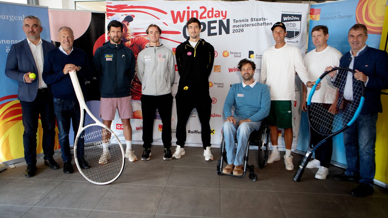 Eine Gruppe von Männern in Sportbekleidung, einer im Rollstuhl, posiert für ein Foto vor einem Schild mit 'Win2day Open'. Sie alle lächeln.