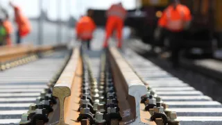 Railway workers in orange suits working on track in a blurry background. Metal rail lines on the foreground with nuts and bolts.