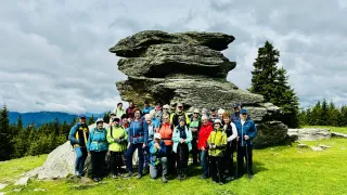 A group of hikers stands in front of a large rock formation, some with hats and sunglasses, holding trekking poles.