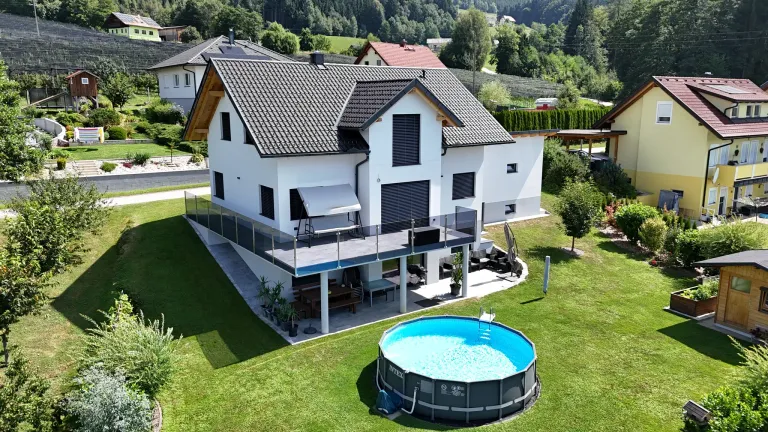 A modern white house with a swimming pool and a balcony is surrounded by greenery and mountains.