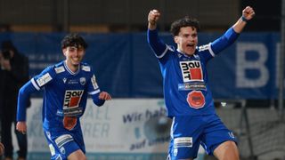 Zwei Fußballer feiern in blauen Trikots, einer mit erhobenen Armen und einem großen Lächeln. Der Spieler vorne trägt ein rotes Logo auf der Brust.