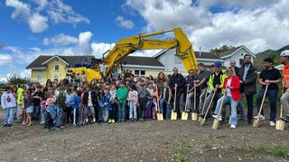 A large crowd of people gather around a yellow excavator, holding shovels, likely participating in a groundbreaking ceremony. In the background, houses are visible under a cloudy sky.