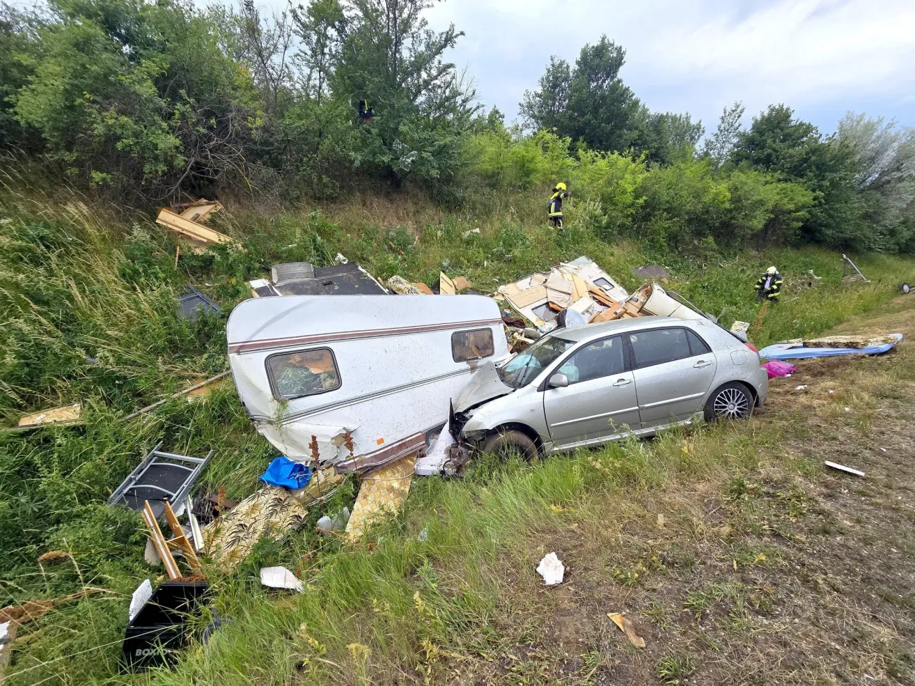 A car and a trailer crashed on a roadside, with debris scattered around. Two people in yellow uniforms are inspecting the scene.