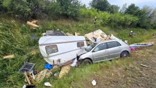 A car and a trailer crashed on a roadside, with debris scattered around. Two people in yellow uniforms are inspecting the scene.