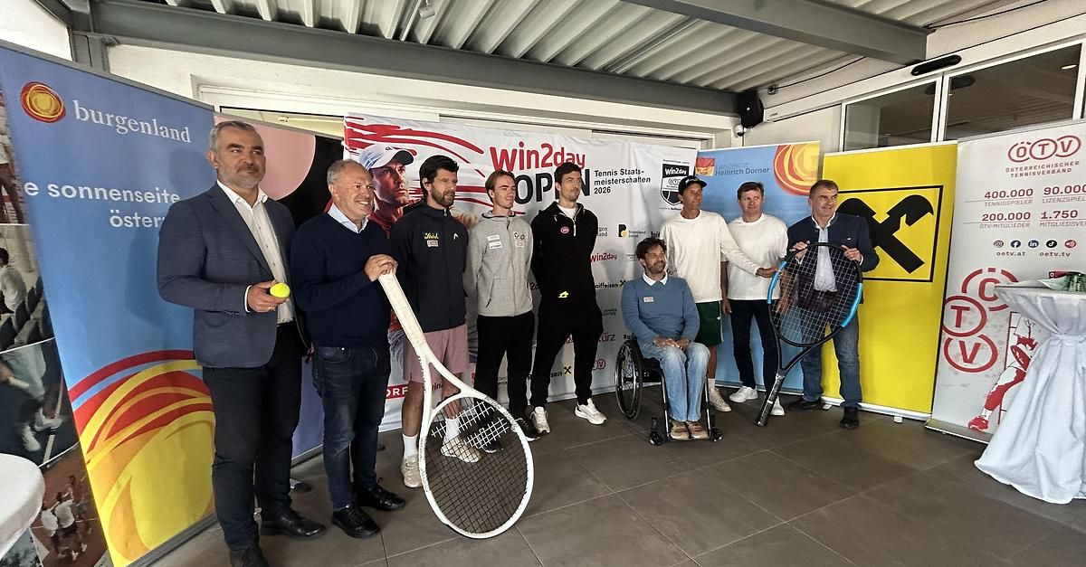 Eine Gruppe von Männern, einige mit Tennisschlägern, steht neben einem Banner, ein Mann im Rollstuhl, wahrscheinlich bei einer Tennisveranstaltung.