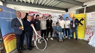 Eine Gruppe von Männern, einige mit Tennisschlägern, steht neben einem Banner, ein Mann im Rollstuhl, wahrscheinlich bei einer Tennisveranstaltung.