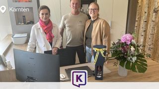 Three individuals stand behind a desk, smiling for a photo. A woman on the left wears a white coat and scarf, a man in the center wears a white shirt with a logo, and a woman on the right wears a beige jacket. A flower vase and monitor are on the desk.