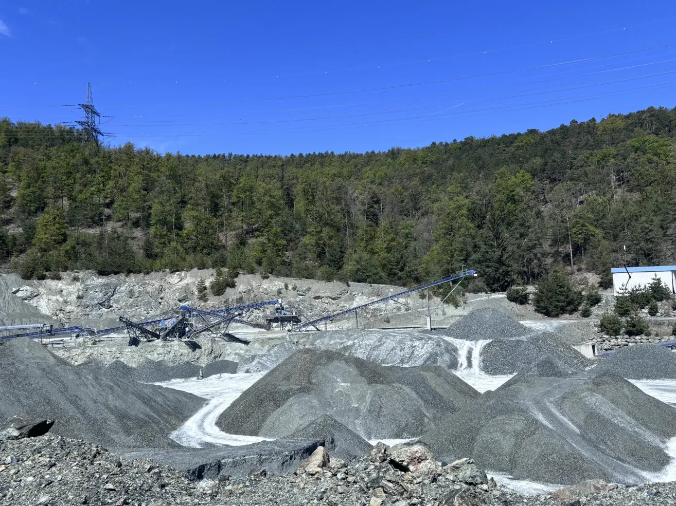 Ein Outdoor-Mining-Standort mit einem blauen Förderband, Kieshaufen und einem bewaldeten Hügel unter einem klaren blauen Himmel.
