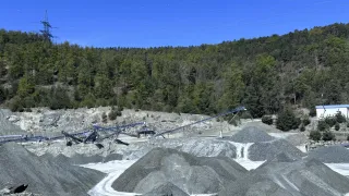 Ein Outdoor-Mining-Standort mit einem blauen Förderband, Kieshaufen und einem bewaldeten Hügel unter einem klaren blauen Himmel.