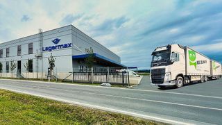 A white truck drives past a large Lagermax warehouse with a fenced area and a blue canopy.