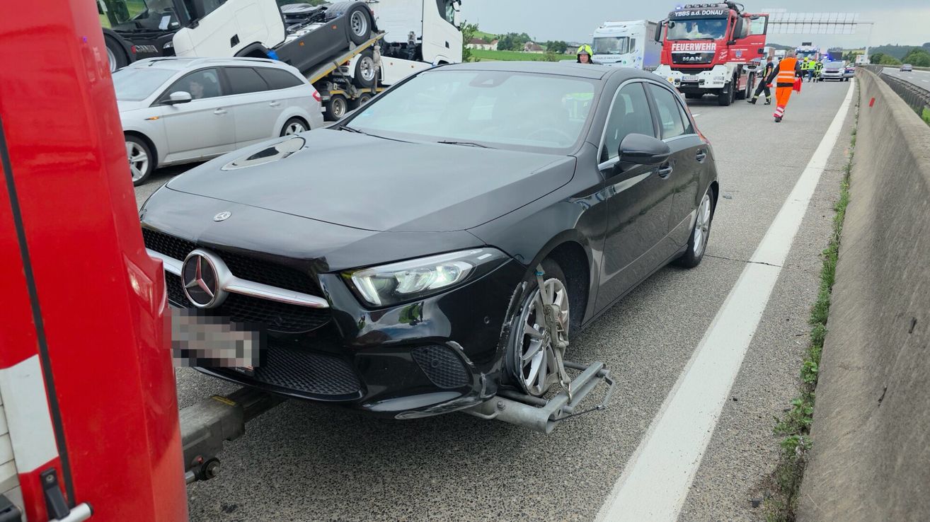 A damaged black Mercedes car is being towed with its front end damaged on the side of the road. Other vehicles, including a truck, are nearby.