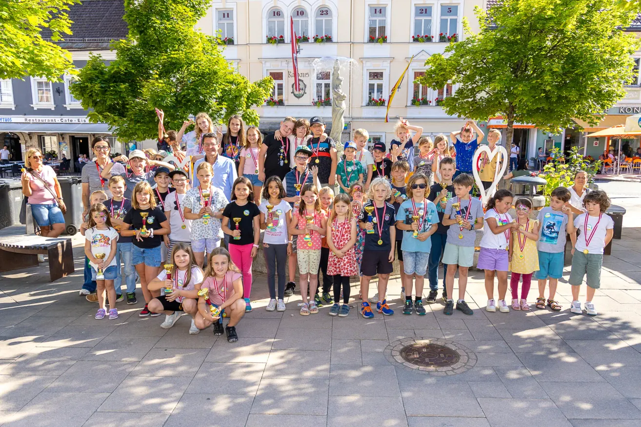 A group of children and adults pose for a photo, all wearing medals around their necks. The scene is outdoors with trees and a building in the background.