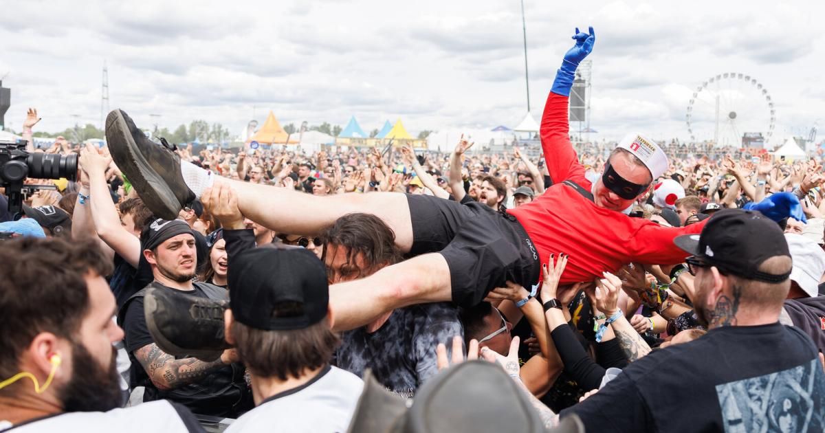 Ein Mann in einem roten Shirt, schwarzen Shorts und blauen Handschuhen crowdsurft bei einem Open-Air-Konzert. Die Menge jubelt und hebt die Hände.