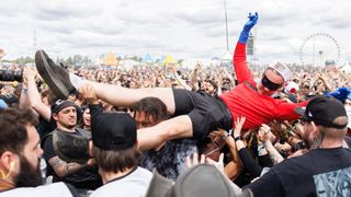 A man wearing a red shirt, black shorts, and blue gloves is crowd surfing at an outdoor concert. The crowd is cheering and raising their hands.