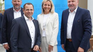 Four individuals are posing for a photo in front of a blue backdrop. A woman in white and three men in dark suits. They appear to be business executives.
