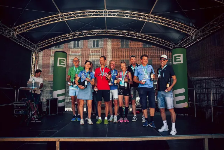 Gewinner eines Events stehen mit Weinflaschen und Medaillen auf der Bühne. Dahinter ein großes Gebäude mit einem Bogenfenster.