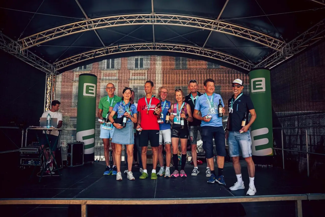 Winners of an event stand on a stage holding wine bottles and medals. Behind them, a large building with an arched window.