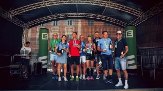 Winners of an event stand on a stage holding wine bottles and medals. Behind them, a large building with an arched window.