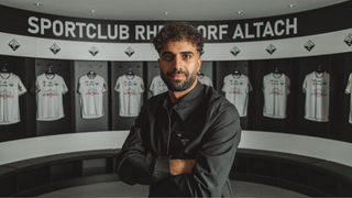 A man stands in front of a locker room with several jerseys hanging, possibly a sports team's uniform. The wall behind him has a banner with the text 'PORTCLUB RH ORF ALTACH.'