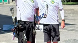 Zwei Männer stehen auf einem roten Teppich, einer hält einen Bogen und Pfeile und der andere einen Bogen. Sie tragen beide weiße T-Shirts und schwarze Shorts. Sie stehen auf einer Straße.