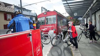 A woman with a bicycle boards a red train at the station. Several people are standing around the train, some with bikes.