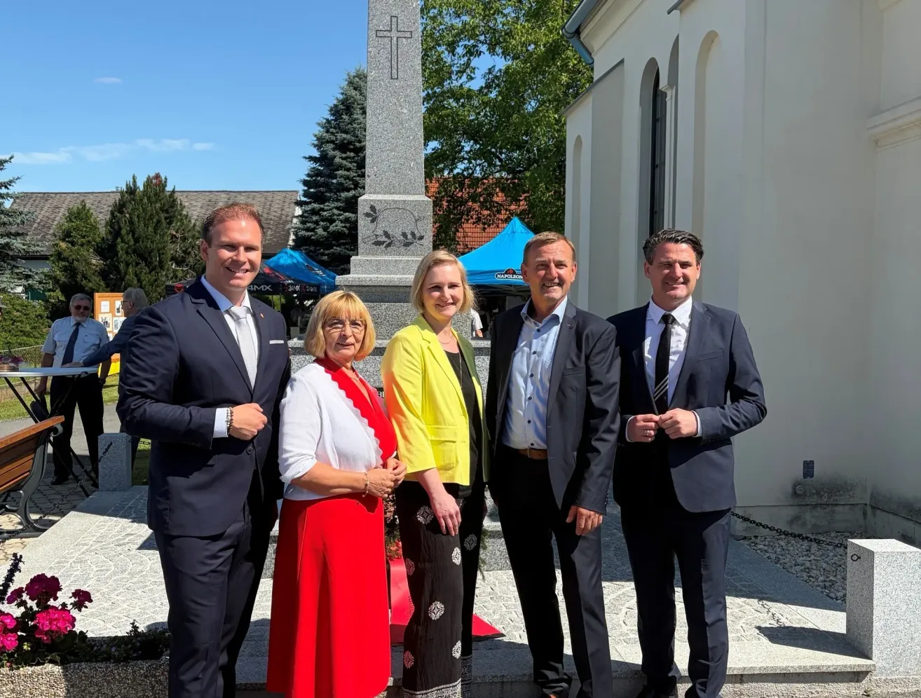 Five people, dressed formally, stand in front of a church and a monument. Behind them are tents and trees.