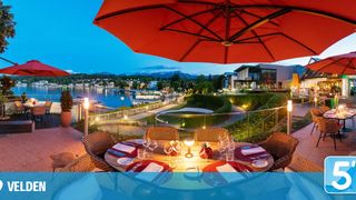 A table set for dinner under a red umbrella, with a view of a lake, mountains, and a golf course.